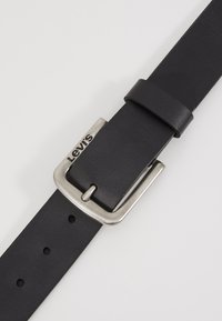 Cinturón de cuero negro con una hebilla de metal plateada. La hebilla presenta un logo grabado de "Levi's", y el cinturón tiene agujeros espaciados uniformemente a lo largo de su longitud.