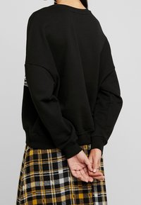 Sweat-shirt noir à manches longues avec poignets côtelés ; présente un design uni et une coupe décontractée, porté avec une jupe à carreaux noire, jaune et blanche.