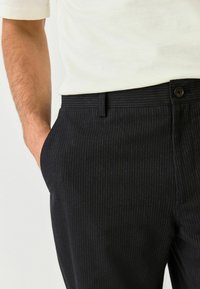 Egy ember felsőteste fehér ingben, sötét csíkos nadrágot viselve, az egyik keze a zsebében pihen, megmutatva egy stílusos öltözéket.