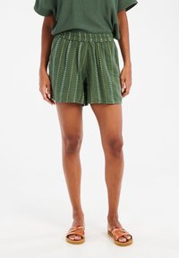 Shorts vert olive avec une taille à cordon de serrage, arborant un motif à rayures verticales dans des teintes plus claires. Associés à des sandales marron à lanières.