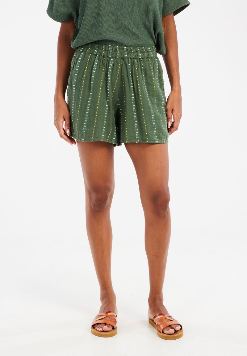 Shorts vert olive avec une taille à cordon de serrage, arborant un motif à rayures verticales dans des teintes plus claires. Associés à des sandales marron à lanières.