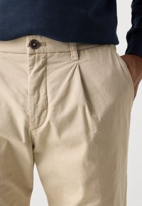 Beige trousers made of lightweight fabric, featuring a button closure, side pockets, and visible pleats for a relaxed fit.