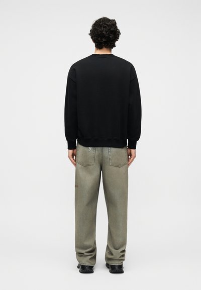 Άτομο με σγουρά σκούρα μαλλιά στέκεται γυρισμένο προς τα πίσω, φορώντας μια μαύρη φούτερ, φαρδιά ξεθωριασμένα γκρι τζιν και μαύρα παπούτσια σε απλό φόντο.