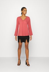 Blusa de manga longa, com decote em V, na cor coral, combinada com uma saia mini preta com uma fenda frontal. Uns sapatos de salto preto com tiras completam o conjunto.