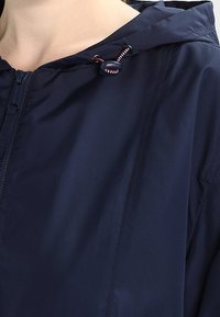 Námořnická modrá bunda s kapucí, s předním zipem, nastavitelnou šňůrkou s červenými a bílými akcenty a hladkým, vodoodpudivým materiálem.