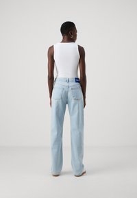 Jeans lejera de culo larg cu talie înaltă, de culoare albastru deschis, având o etichetă albastră pe spate și două buzunare posterior. Purtați cu un top alb.