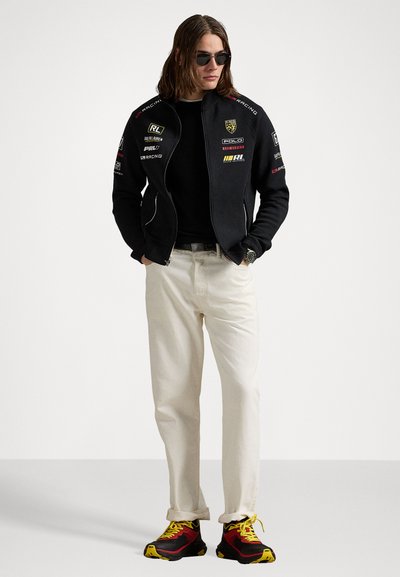 Schwarze Zip-Jacke mit Rennlogos, schwarzes Shirt, hellbeige Hose und bunte Sneakers mit roten, gelben und schwarzen Akzenten.
