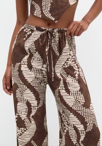 Pantaloni marroni con motivo, realizzati in materiale leggero, con vita regolabile tramite coulisse. Presentano forme geometriche bianche e un design fluido.