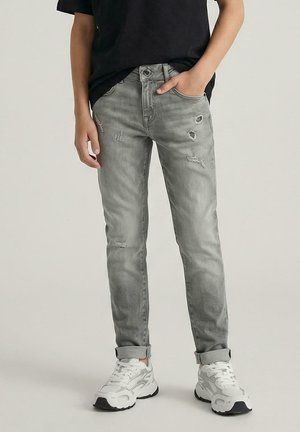 Kind draagt lichtgrijze gebleekte jeans opgerold bij de pijpen, witte sneakers en een zwart shirt met één hand in de zak.