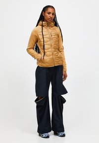 Pufferjack in goudbeige met een ritssluiting aan de voorkant en een capuchon; gecombineerd met donkerblauwe wijde broek met splitten aan de zomen en een gestructureerde stof.