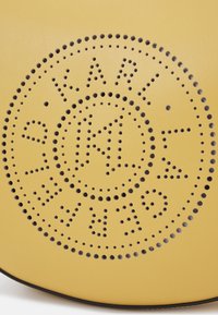 Borsa rotonda in pelle gialla con un design perforato con il logo "KARL" e accenti circolari. Texture liscia con perforazioni scure a contrasto.