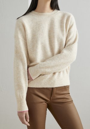 Cream-colored knit sweater with a round neckline and pink-accented cuffs, paired with brown pants. Soft texture and relaxed fit.