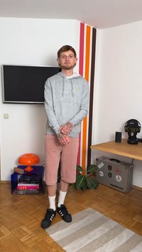 Hoodie cinza com bolso canguru, calções de veludo-cotelê rosa, meias brancas e sapatilhas negras. O fundo apresenta uma planta, móveis e riscas.