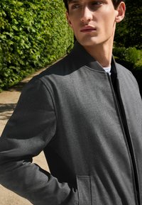 Graue Bomberjacke mit glatter Textur, durchgehendem Reißverschluss und seitlichen Taschen. Verfügt über einen Stehkragen und ein minimalistisches Design, vor einer grünen Kulisse.