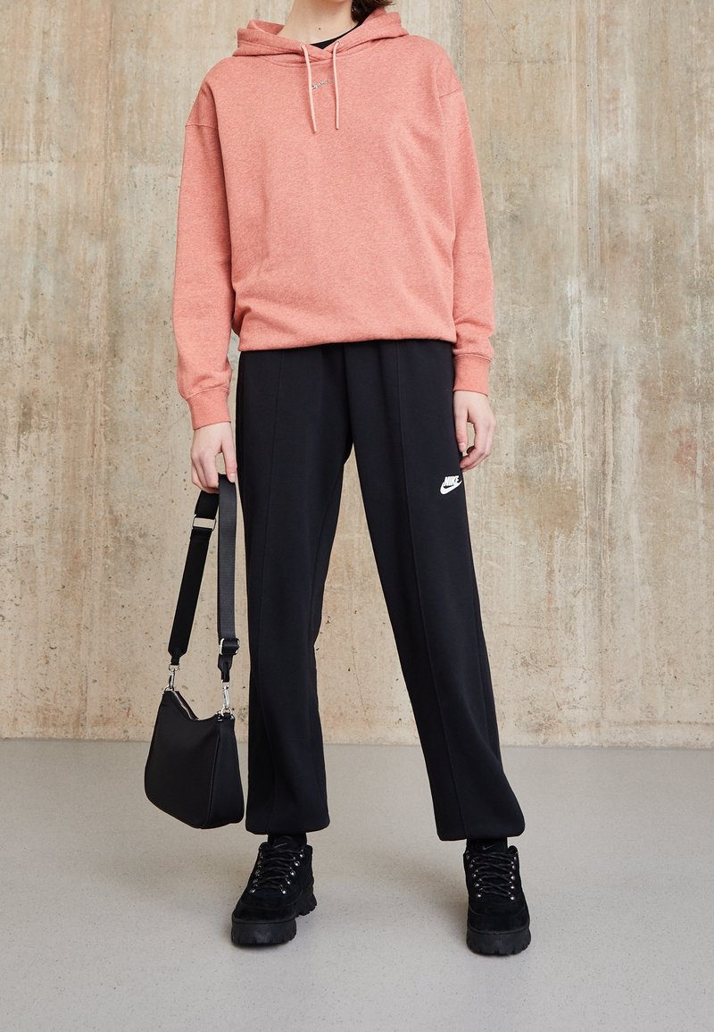 Personne portant un sweat à capuche rose, un pantalon de survêtement Nike noir, des baskets noires, tenant un sac à bandoulière noir devant un mur en béton.