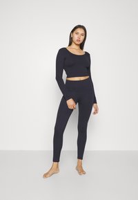 Marinblå, ribbad långärmad topp och matchande leggings. Stretchigt tyg, figurnära design, rundad halsringning. Modell stående barfota mot en vit bakgrund.