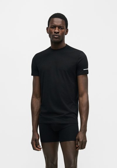 Άνδρας που φοράει στενή μαύρη μπλούζα με κοντά μανίκια και μαύρα σορτς που ταιριάζουν, στέκεται μπροστά σε απλό φωτεινό φόντο.