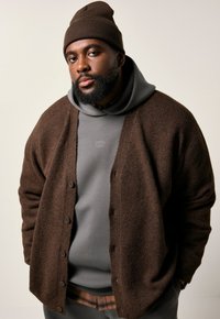 Bruntexturerad cardigan över en grå hoodie, som har en logotyp, tillsammans med en matchande brun stickad mössa. Outfiten har en avslappnad passform och knapprading.