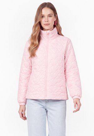 Jeune femme portant une veste matelassée rose clair avec des motifs de cœurs et un jean bleu clair, debout devant un fond blanc uni.