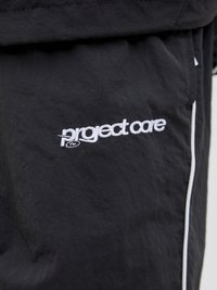 Sorte stoffbukser med en subtil tekstur, hvit "project care"-logo og kontrasterende hvite sideaksenter langs sømmene.