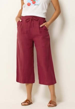 Kobieta nosząca wysokie, szerokie spodnie 7/8 w kolorze bordowym z paskiem ściągającym i bocznymi kieszeniami, w połączeniu z brązowymi sandałami na płaskim obcasie i białym topem.