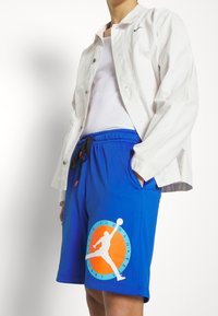 Shorts atletici blu con texture in rete e un logo arancione raffigurante una silhouette che salta. Accompagnati da una camicia bianca con bottoni e una canotta.