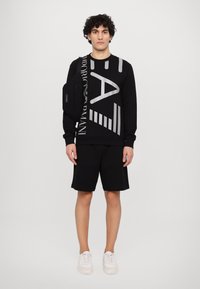 Felpa nera con grande scritta grafica grigia, abbinata a pantaloni corti neri. Sneakers bianche e una piccola borsa a tracolla nera.