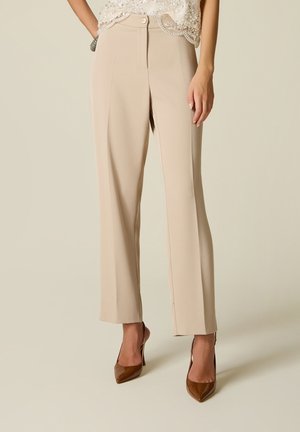 Beige, hoog getailleerde pantalon gedragen met bruine puntschoenen en een witte versierde top tegen een lichte achtergrond.