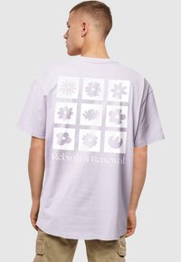 Lilla bomulds t-shirt med et stort, gitterlignende blomstermotiv i forskellige nuancer af lilla med teksten "Genfødsel & Fornyelse" nedenfor.