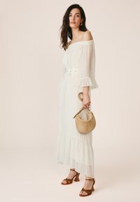 Femme en robe blanche épaules dénudées avec manches et ourlet à volants, tenant un sac à main beige en forme de croissant, portant des sandales marron à talons.