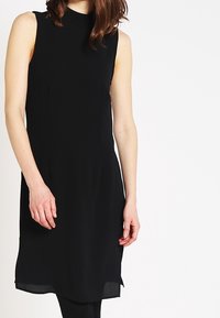 Svart ärmlös klänning med hög hals, tillverkad av slätt tyg. Har en rak skärning och något längre nederkant bak. Inga mönster eller detaljer.