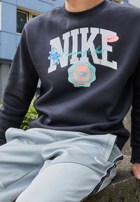 Hombre vistiendo un sudadera negra de Nike con un logo colorido y pantalones grises claros de Nike, sentado al aire libre cerca de un edificio y vegetación.