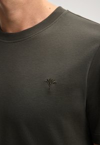 Dunkelgrünes T-Shirt mit Rundhalsausschnitt, glatter Textur und einem kleinen gestickten Blumenmuster in einem passenden Grünton auf der Brust.