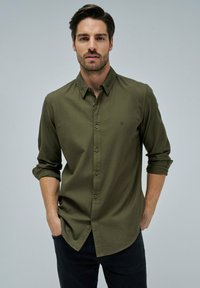 Camicia button-up a quadri verde oliva con maniche lunghe, arrotolate ai polsini. Presenta un pattina frontale nascosta e un piccolo dettaglio del logo.