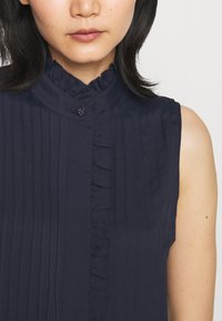 Námořnicky modrá halenka bez rukávů s vertikálními tenkými pruhy, volánkovým límcem a zapínáním na knoflíky, vyrobená z lehké látky.