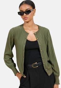 Olivgrüner gerippter Cardigan mit rundem Halsausschnitt, vorderem Knopfverschluss und langen Ärmeln. Über einem schwarzen Tanktop und schwarzen Hosen getragen.