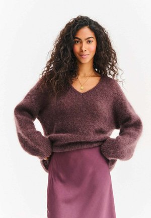 Bordowy puszysty sweter z dekoltem w serek i długimi, szerokimi rękawami, w połączeniu z gładką fioletową satynową spódnicą, na białym tle.