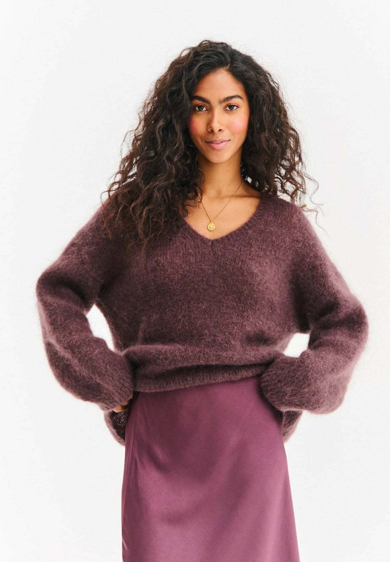 Bordowy puszysty sweter z dekoltem w serek i długimi, szerokimi rękawami, w połączeniu z gładką fioletową satynową spódnicą, na białym tle.