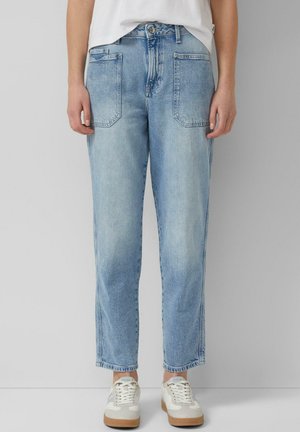 Osoba ubrana w jasnoniebieskie jeansy z wysokim stanem i przednimi kieszeniami naszywanymi oraz białe trampki, stojąca na tle jednolitego jasnego tła.