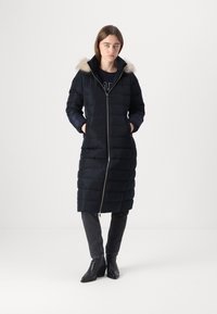 Piumino nero con design a lunghezza media, cappuccio foderato in pelliccia e chiusura frontale con zip. Presenta una texture trapuntata e tasche laterali.