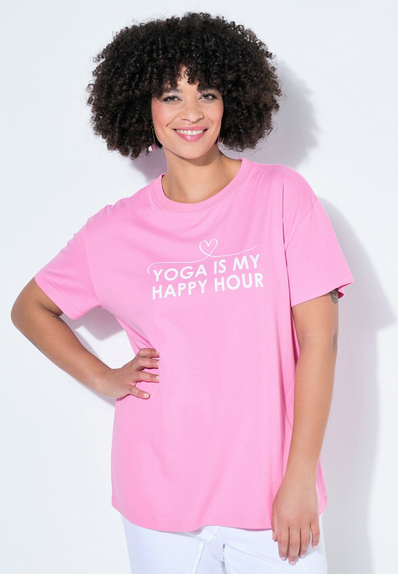 Frau mit lockigem Haar, die lächelt und ein rosa T-Shirt mit der Aufschrift „YOGA IST MEINE GLÜCKSSTUNDE“ trägt, steht mit einer Hand in der Hüfte vor weißem Hintergrund.