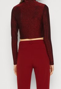Gekürztes langärmliges Oberteil aus rotem, glitzerndem Material; hat einen hohen Kragen und ein figurbetontes Design. Die Hose ist hoch geschnitten in einfarbigem Rot.