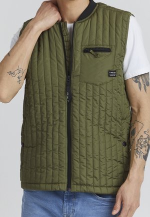 Homme portant une veste sans manches matelassée vert olive avec fermeture éclair frontale, sur un t-shirt blanc et un jean bleu, montrant des tatouages sur l'avant-bras.