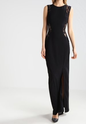 Frau trägt ein ärmelloses schwarzes Abendkleid mit seitlichen Spitzendetails und einem Frontschlitz, kombiniert mit schwarzen spitzen Schuhen.