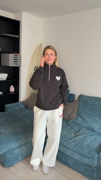 Γυναίκα που χαμογελάει, φορώντας μαύρο φούτερ και άσπρο παντελόνι, στέκεται κοντά σε μπλε καναπέ και σανίδα του σερφ σε ένα φωτεινό σαλόνι με ράφια.