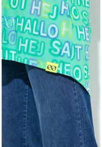 Grøn skjorte med flerfarvede "hej"-ord på forskellige sprog over blå jeans, med en lille gul etiket med et sort uendelighedssymbol.