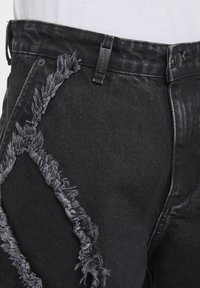 Jean en denim noir avec des détails effilochés le long de la poche avant et de la cuisse, porté avec une chemise blanche rentrée.