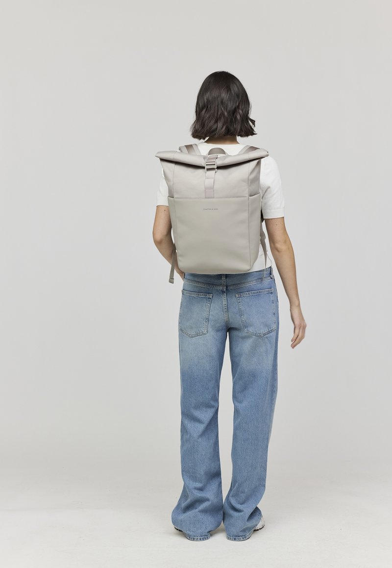 Sac à dos roll-top gris clair avec des bretelles réglables et une boucle à l'avant. Texture lisse, design minimaliste et petit logo de la marque au dos.