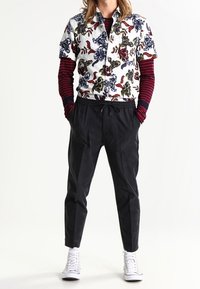 Camicia bianca a maniche corte con un motivo floreale colorato, abbinata a pantaloni neri con coulisse e sneakers bianche. Sotto, una maglietta a maniche lunghe a righe.