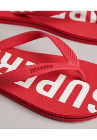 Tongs en caoutchouc rouge avec une surface texturée. Présentent une lanière en chevrons et le texte blanc "SUPERDRY" sur la semelle.